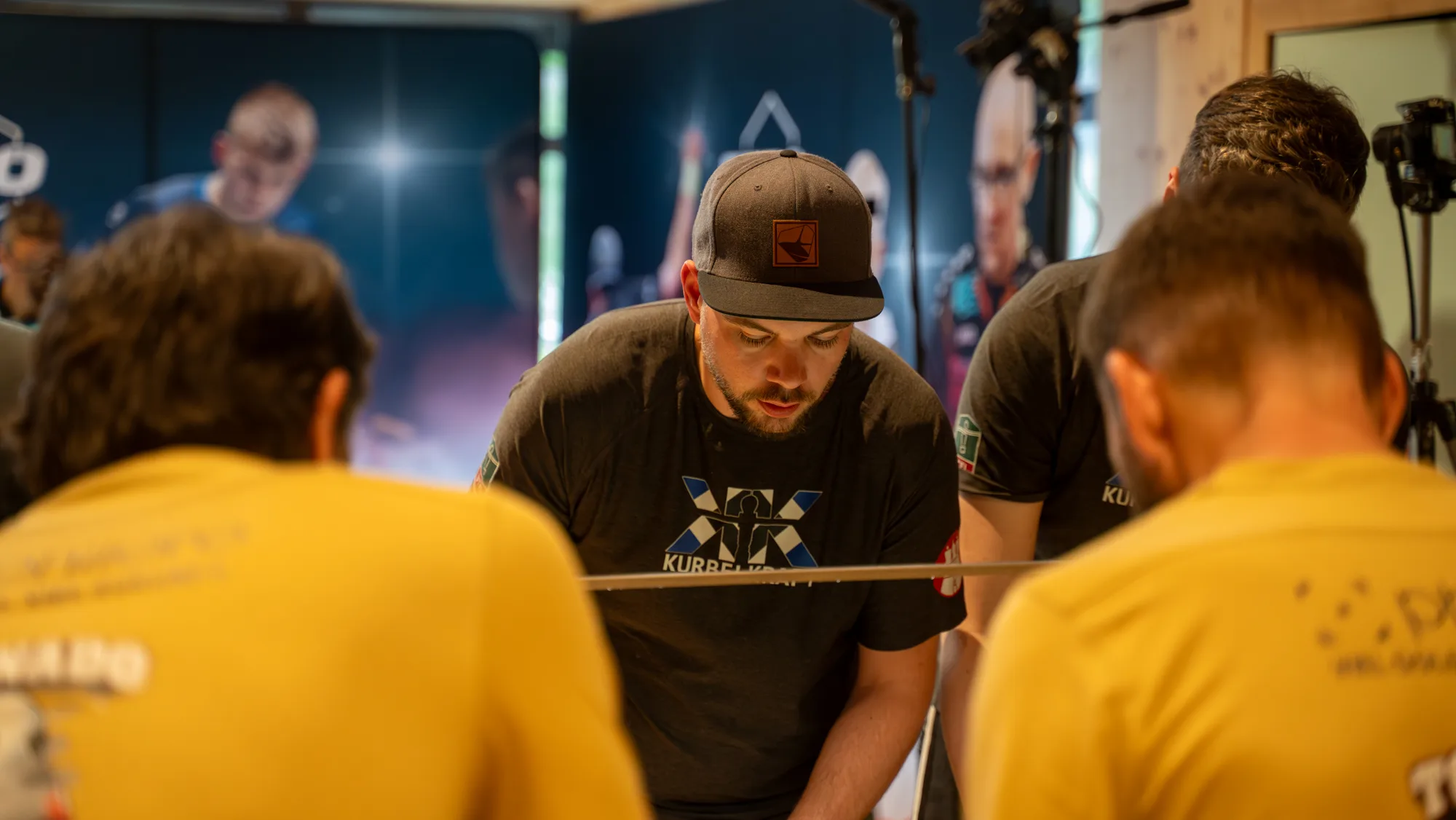 Mario Christ mit Kurbelkraft-Cap – hochkonzentriert über dem Kicker-Tisch, Gegner verschwommen im Vordergrund, Turnier-Atmosphäre