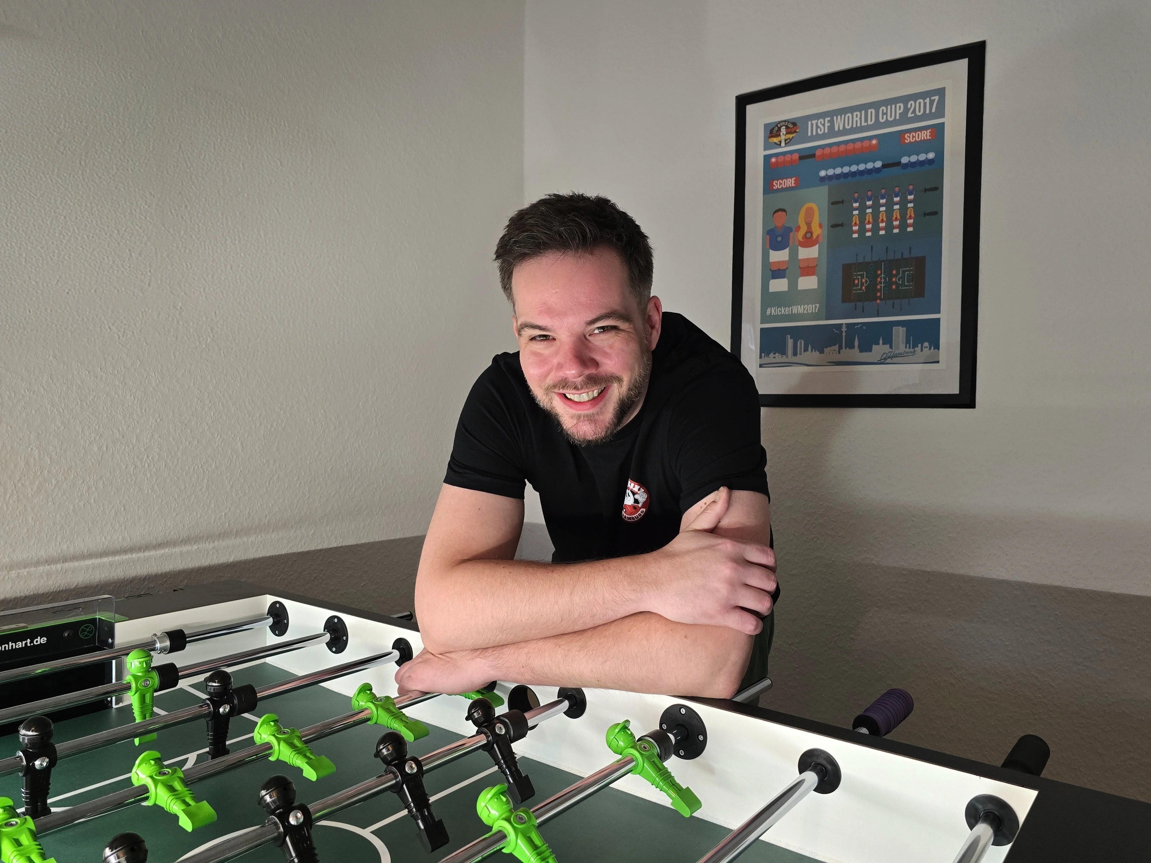 Mario Christ am Kicker-Tisch, lachend, ITSF World Cup Poster im Hintergrund